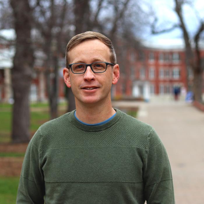 Zach Whalen standing in the foreground, behind him a tree-lined path to a building